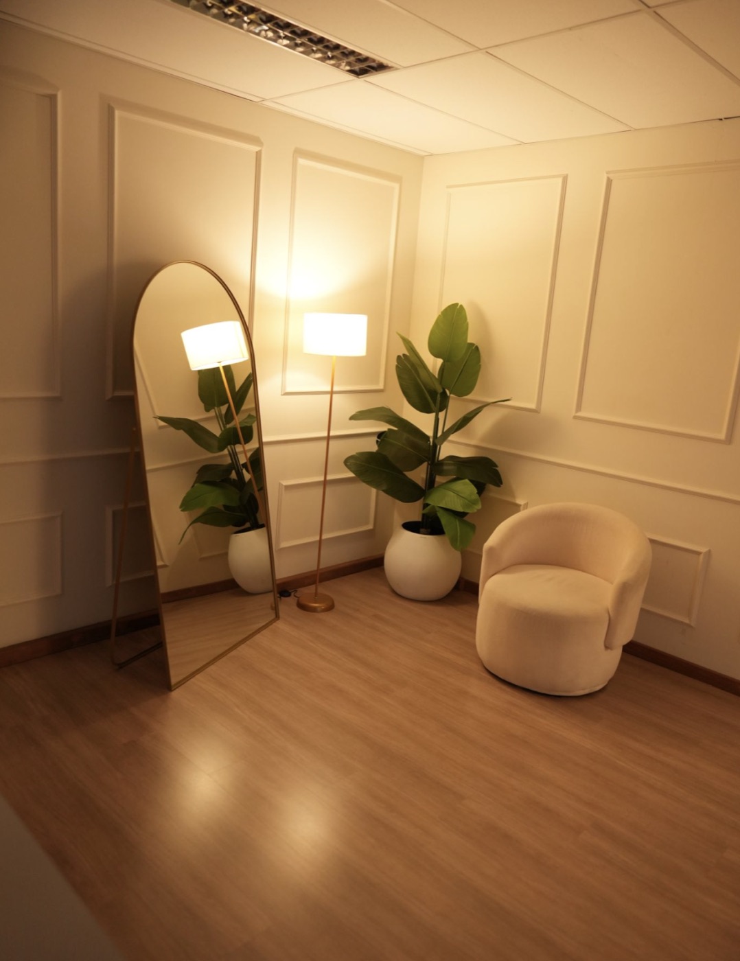 Cenário com parede boiserie branca, espelho arco, poltrona bouclé e plantas